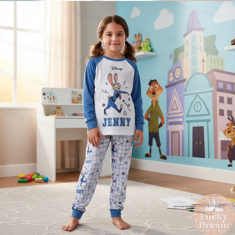Peut inclure: Une jeune fille porte un ensemble de pyjama Disney. Le haut est blanc avec des manches bleues et pr&eacute;sente un dessin de Judy Hopps et le nom "JENNY". Le pantalon de pyjama est bleu clair avec un motif &agrave; carreaux et des graphiques de Judy Hopps.
