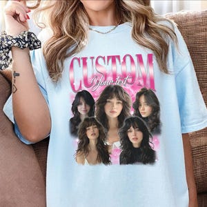 May include: Light blue t-shirt with a custom design. The design features the word "CUSTOM" in pink, with the words "Your text" below it. Below the text are five images of women with long hair.