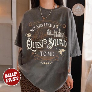 Op de afbeelding: Een grijs T-shirt met een vintage-stijl grafisch ontwerp met een kasteel, draken en de tekst "Sounds Like A Quest Squad To Me".