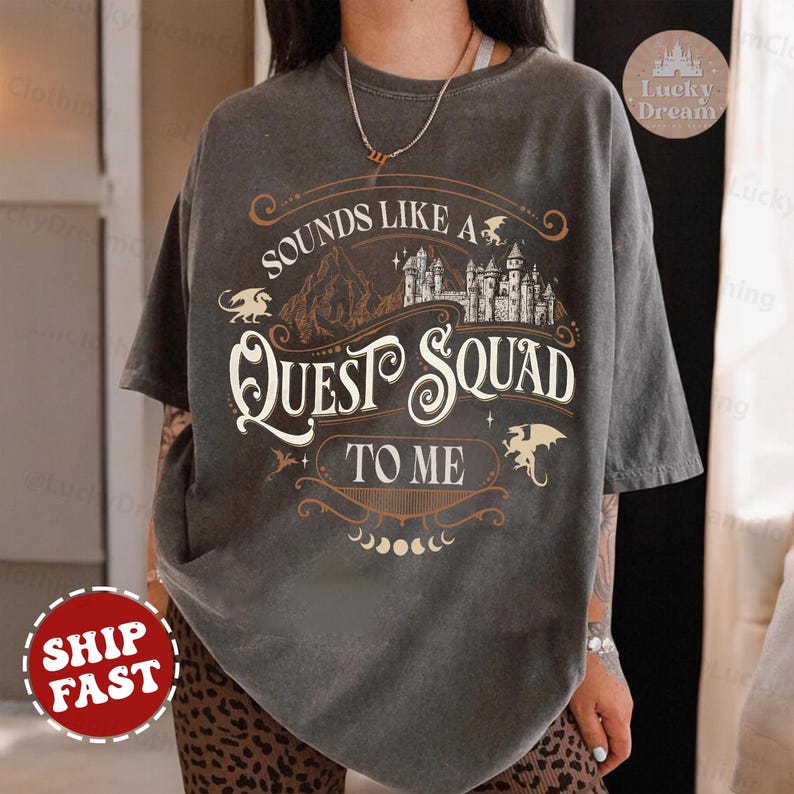 Op de afbeelding: Een grijs T-shirt met een vintage-stijl grafisch ontwerp met een kasteel, draken en de tekst "Sounds Like A Quest Squad To Me".