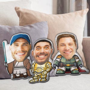 Puede incluir: Tres cojines personalizados con caras sonrientes, cada uno con un disfraz de personaje de Star Wars diferente. Uno tiene un sable de luz azul, otro es dorado y el tercero es verde y marr&oacute;n. En un sof&aacute;.