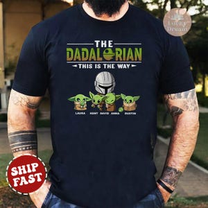 May include: A black t-shirt with a graphic of a Mandalorian character and four Baby Yoda characters. The text "The Dadalorian - This is the Way" is printed on the shirt. The names Laura, Kent, David, Anna, and Dustin are printed below the characters.