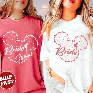 Può includere: Due magliette bianche con un design a forma di orecchio di Topolino rosso. Una maglietta dice "Bride" e l'altra dice "Bride Squad".