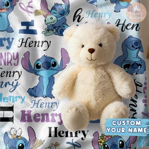 Puede incluir: Un oso de peluche blanco se sienta sobre una manta blanca y azul con un patr&oacute;n repetido de un personaje de dibujos animados azul con orejas grandes y el nombre "Henry" repetido en toda la manta.