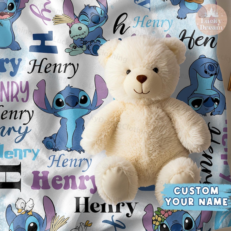 Puede incluir: Un oso de peluche blanco se sienta sobre una manta blanca y azul con un patr&oacute;n repetido de un personaje de dibujos animados azul con orejas grandes y el nombre "Henry" repetido en toda la manta.