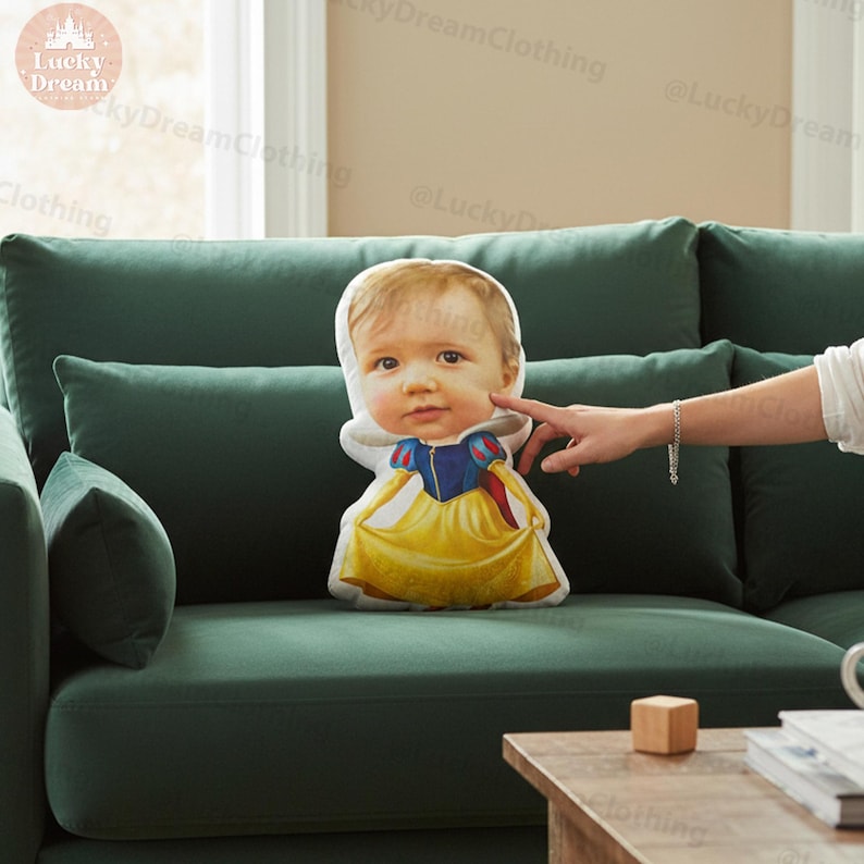 May include: A custom-printed pillow shaped like a child dressed as Snow White, featuring a photo of a baby's face. The pillow has a white face, a blue top, and a yellow dress. The pillow is on a green couch.