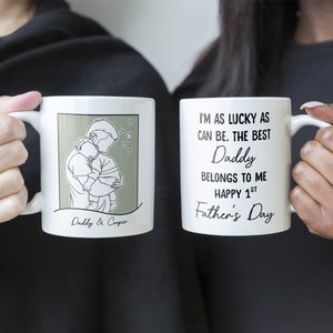 K&ouml;nnte beinhalten: Zwei wei&szlig;e Tassen mit schwarzem Text und Illustrationen. Eine Tasse zeigt eine Strichzeichnung eines Vaters, der ein Baby h&auml;lt, mit dem Text "Daddy & Cooper". Die andere Tasse hat den Text "Ich bin so gl&uuml;cklich wie es geht. Der beste Papa geh&ouml;rt mir. Frohen 1. Vatertag."