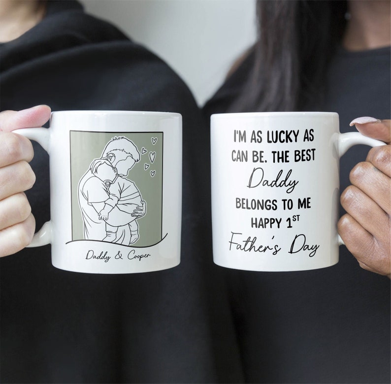 K&ouml;nnte beinhalten: Zwei wei&szlig;e Tassen mit schwarzem Text und Illustrationen. Eine Tasse zeigt eine Strichzeichnung eines Vaters, der ein Baby h&auml;lt, mit dem Text "Daddy & Cooper". Die andere Tasse hat den Text "Ich bin so gl&uuml;cklich wie es geht. Der beste Papa geh&ouml;rt mir. Frohen 1. Vatertag."