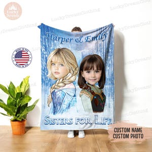Op de afbeelding: Een blauwe en witte fleece deken met een aangepaste foto van twee meisjes die verkleed zijn als Elsa en Anna uit Frozen. De tekst "Harper & Emily Sisters for Life" staat op de deken gedrukt. De deken is gemaakt in de VS.