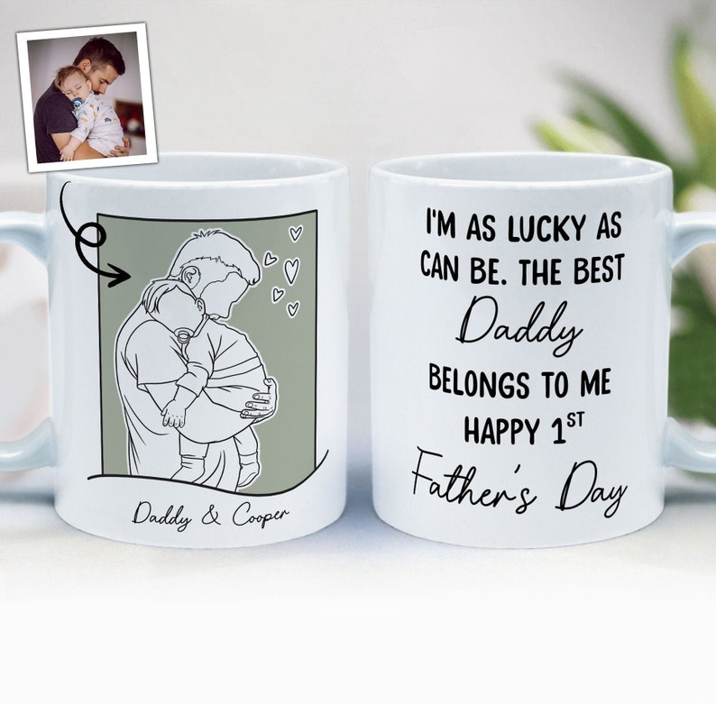 K&ouml;nnte beinhalten: Wei&szlig;e Keramiktasse mit einer Schwarzwei&szlig;-Zeichnung eines Vaters, der ein Kind h&auml;lt. Die Tasse hat den Text "Daddy & Cooper" auf der Unterseite und "I'm as lucky as can be. The best Daddy belongs to me. Happy 1st Father's Day" auf der anderen Seite.