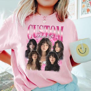 May include: Pink sweatshirt with a graphic design featuring the word "CUSTOM" in a pink gradient font, with the words "Your text" below. The design includes five images of a woman with long hair. A white mug with a yellow smiley face is held in the hand.