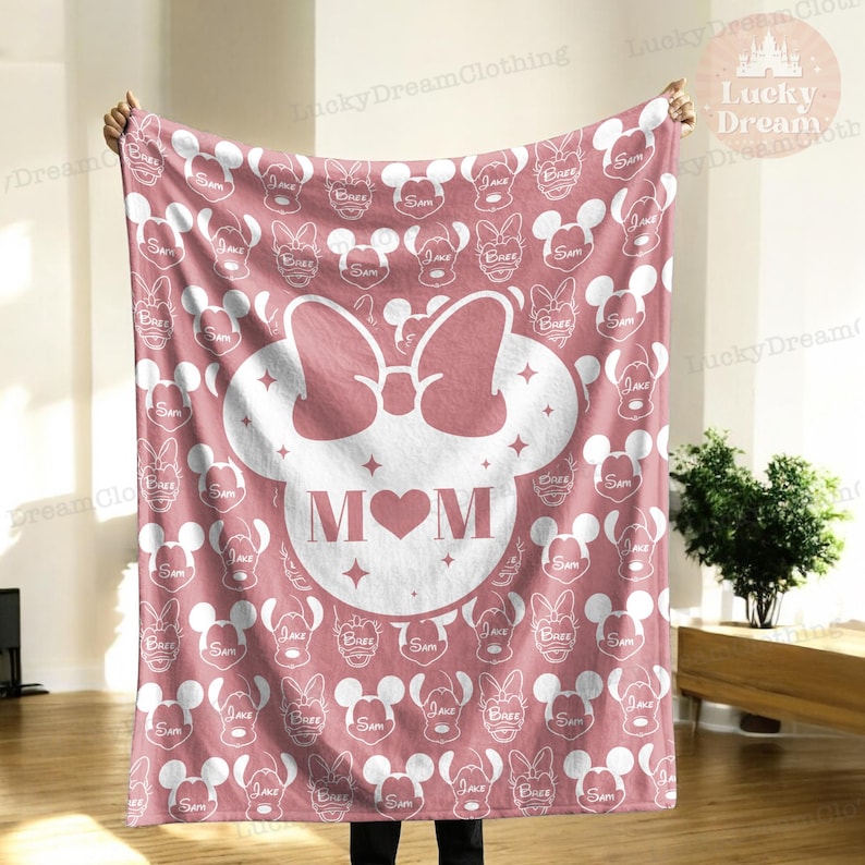 K&ouml;nnte beinhalten: Eine rosa Fleece-Decke mit einem wei&szlig;en Mickey-Maus-Kopf-Design. Der Kopf hat eine rosa Schleife und das Wort "MOM" mit einem rosa Herzen in der Mitte. Der Hintergrund ist mit einem sich wiederholenden Muster aus wei&szlig;en Mickey-Maus-K&ouml;pfen bedeckt.