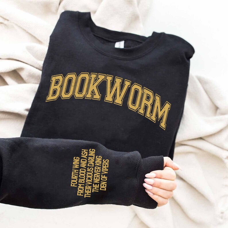 Pode incluir: Camisola de moletom preta com o texto "BOOK WORM" em letras douradas na frente. A manga tem o texto "FOURTH WING", "FROM BLOOD AND ASH", "THEIR VICIOUS DARLING", "THE NEVER KING" e "DEN OF VIPERS" em letras douradas.
