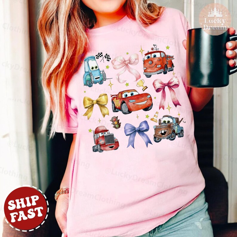 May include: Pink t-shirt with a colorful cartoon design featuring cars from the Disney Pixar movie Cars. The design includes Lightning McQueen, Mater, and other characters. The shirt has a bow tie design.