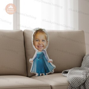 May include: A custom-made pillow featuring a smiling child's face, dressed in a blue gown with a cape. The pillow is shaped like the child and is placed on a beige sofa. The pillow is a unique personalized gift.