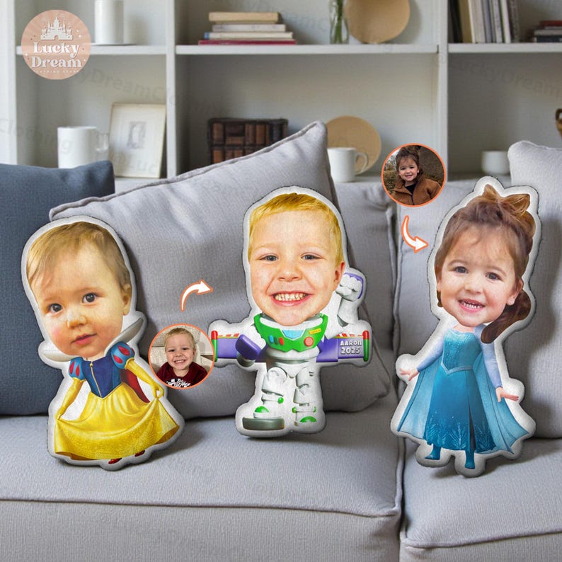 May include: Three custom-made plush pillows featuring children's faces. One pillow is designed as Snow White in a yellow dress, another as Buzz Lightyear, and the third as Elsa in a blue dress. The pillows are on a gray couch.