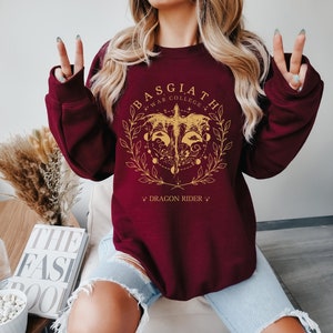 May include: Burgundy sweatshirt with a gold graphic of a dragon with the text "Basgiath War College" and "Dragon Rider" on a maroon background.