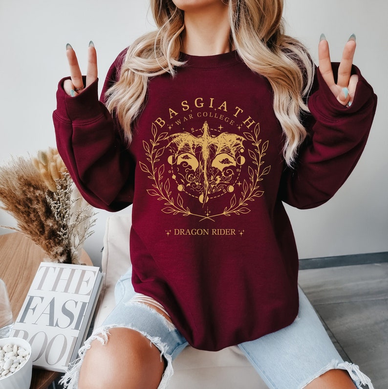 May include: Burgundy sweatshirt with a gold graphic of a dragon with the text "Basgiath War College" and "Dragon Rider" on a maroon background.