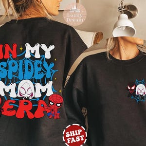 May include: Black sweatshirt with the text "In My Spidey Mom Era" in colorful letters and a spider web design. There are three cartoon spider-man characters on the front of the shirt.