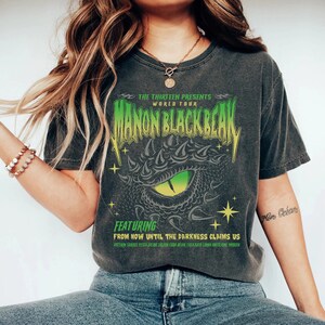 May include: A black t-shirt with a green and black graphic of a dragon's eye. The text "THE THIRTEEN PRESENTS WORLD TOUR MANON BLACKBEAK" is printed on the shirt. The text "FEATURING FROM NOW UNTIL THE DARKNESS CLAIMS US" is printed below the graphic. The text "ASTERIN SORREI VESTA FALINE FALIAN EDSA BEJAR THEA KAYA LINNA BHISLAINE IMBEEN" is printed below the text.