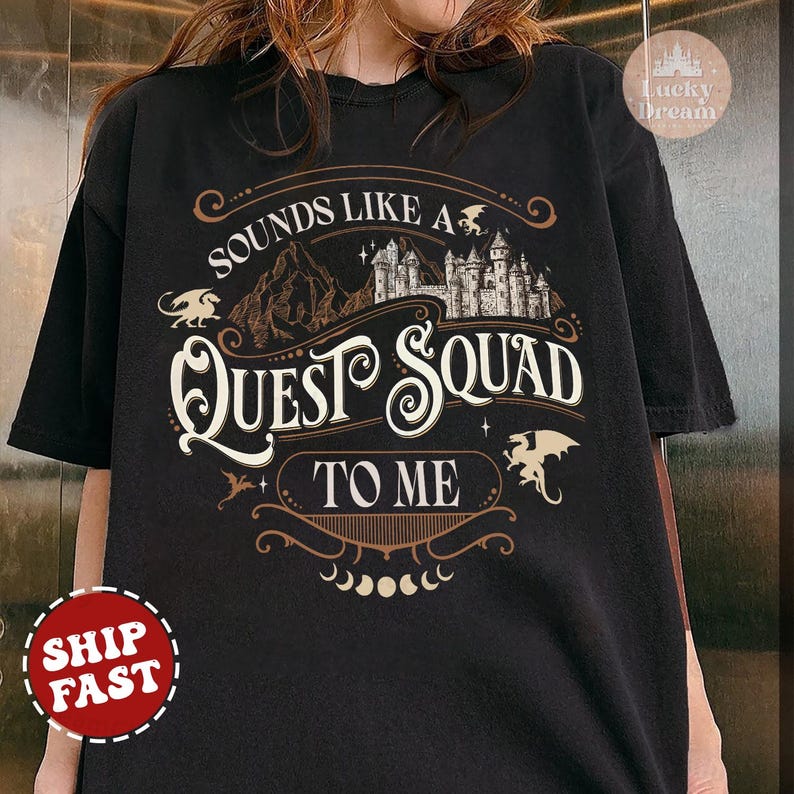 Op de afbeelding: Zwart T-shirt met een bruin grafisch ontwerp met de tekst "Sounds like a Quest Squad to me". De afbeelding bevat een kasteel, bergen en draken.