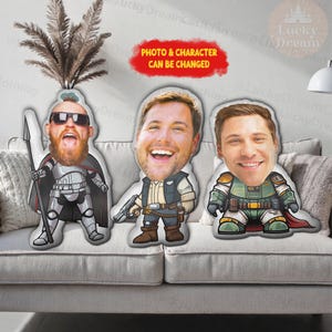 Puede incluir: Tres cojines de personajes personalizados con caras sonrientes, cada uno con un disfraz &uacute;nico. Los cojines est&aacute;n sobre un sof&aacute; gris. El texto "PHOTO & CHARACTER CAN BE CHANGED" est&aacute; en una burbuja roja.