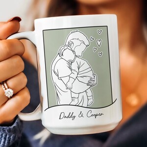 K&ouml;nnte beinhalten: Wei&szlig;e Keramiktasse mit gr&uuml;nem Hintergrund und einer Strichzeichnung eines Vaters, der ein Baby h&auml;lt. Der Text "Daddy & Cooper" ist unter dem Bild gedruckt.