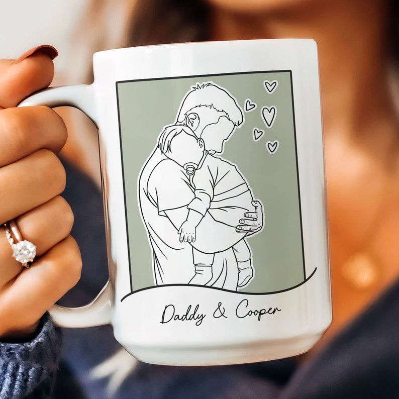 K&ouml;nnte beinhalten: Wei&szlig;e Keramiktasse mit gr&uuml;nem Hintergrund und einer Strichzeichnung eines Vaters, der ein Baby h&auml;lt. Der Text "Daddy & Cooper" ist unter dem Bild gedruckt.