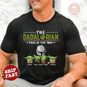 May include: Black t-shirt with a green and yellow graphic of a Mandalorian character and four baby Yoda characters. The text "The Dadalorian This is the Way" is printed on the shirt. The names Laura, Kent, David, Anna, and Dustin are printed below the characters.