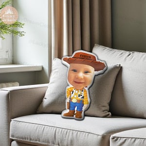 K&ouml;nnte beinhalten: Ein kissenf&ouml;rmiges Kissen mit dem Gesicht eines Babys, das einen Cowboyhut mit dem Namen "Evelyn" tr&auml;gt. Das Baby tr&auml;gt ein Woody-inspiriertes Outfit mit gelbem Hemd, blauer Hose und braunen Stiefeln. Das Kissen liegt auf einem grauen Sofa.