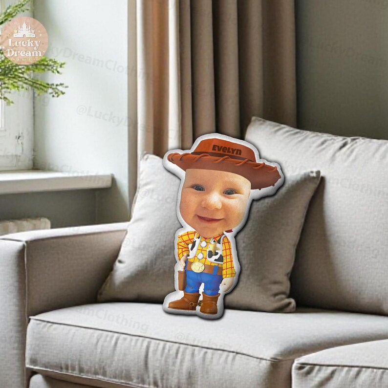 K&ouml;nnte beinhalten: Ein kissenf&ouml;rmiges Kissen mit dem Gesicht eines Babys, das einen Cowboyhut mit dem Namen "Evelyn" tr&auml;gt. Das Baby tr&auml;gt ein Woody-inspiriertes Outfit mit gelbem Hemd, blauer Hose und braunen Stiefeln. Das Kissen liegt auf einem grauen Sofa.