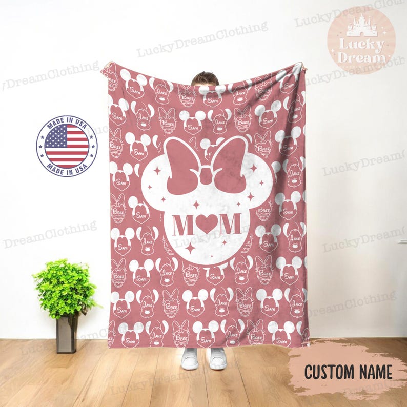 K&ouml;nnte beinhalten: Eine rosa Fleece-Decke mit einer wei&szlig;en Umrandung von Mickey-Maus-Ohren und einer Schleife. Die Ohren enthalten ein Herz mit dem Wort "MOM" darin. Der Hintergrund ist ein rosa Muster aus Mickey-Maus-K&ouml;pfen.
