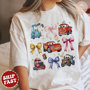 May include: White t-shirt with a colorful cartoon design featuring various cars from the Disney Pixar movie Cars. The design includes Lightning McQueen, Mater, and other characters. The shirt has a crew neck and short sleeves.