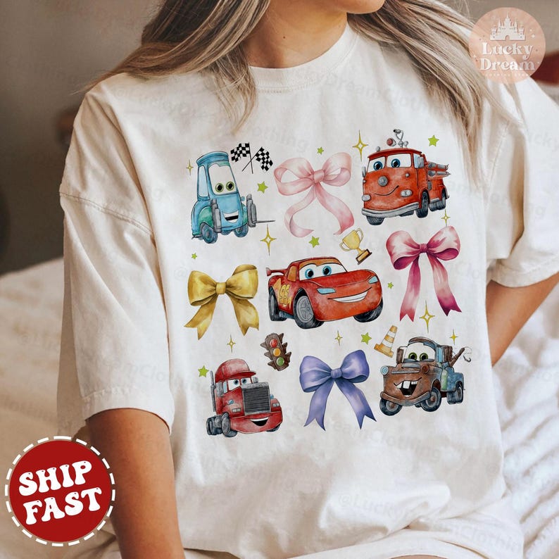 May include: White t-shirt with a colorful cartoon design featuring various cars from the Disney Pixar movie Cars. The design includes Lightning McQueen, Mater, and other characters. The shirt has a crew neck and short sleeves.