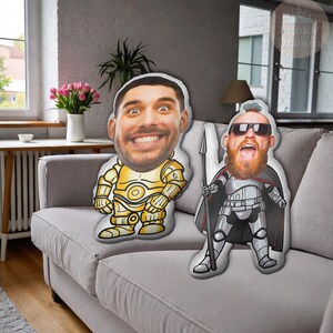 Puede incluir: Dos cojines personalizados con caras impresas. Un coj&iacute;n presenta a una persona con un disfraz de robot dorado, y el otro es una persona con un disfraz de guerrero plateado y negro. Ambos cojines est&aacute;n sobre un sof&aacute; gris.