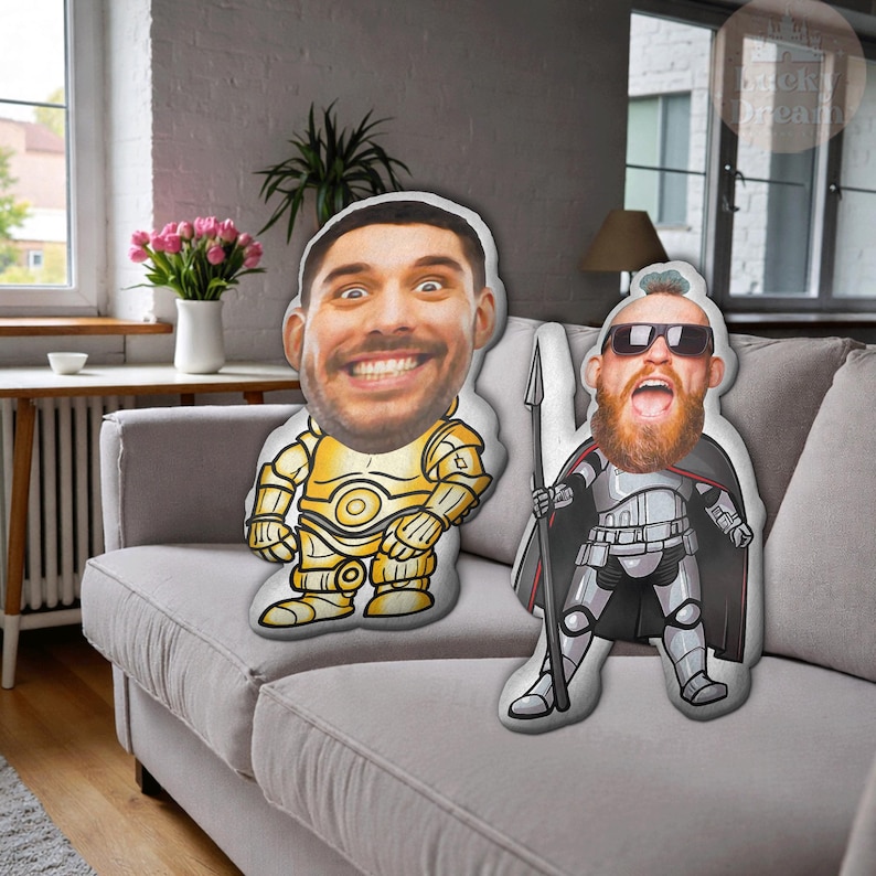 Puede incluir: Dos cojines personalizados con caras impresas. Un coj&iacute;n presenta a una persona con un disfraz de robot dorado, y el otro es una persona con un disfraz de guerrero plateado y negro. Ambos cojines est&aacute;n sobre un sof&aacute; gris.