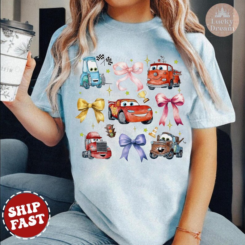 May include: A light blue t-shirt with a cartoon design featuring various cars from the Disney Pixar movie Cars. The cars are surrounded by colorful bows. The shirt has a crew neck and short sleeves.