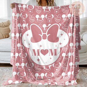 K&ouml;nnte beinhalten: Eine rosa Fleece-Decke mit einer wei&szlig;en Umrandung von Mickey Mouse-Ohren und einer Schleife. Das Wort "MOM" befindet sich innerhalb der Ohren mit einem rosa Herzen in der Mitte. Der Hintergrund ist ein rosa-wei&szlig;es Muster aus Mickey Mouse-K&ouml;pfen mit den Namen "Sam", "Jake" und "Bree" darauf.