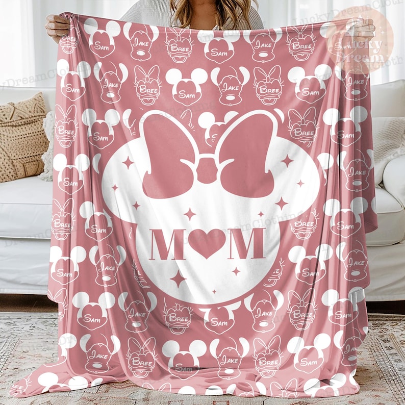 K&ouml;nnte beinhalten: Eine rosa Fleece-Decke mit einer wei&szlig;en Umrandung von Mickey Mouse-Ohren und einer Schleife. Das Wort "MOM" befindet sich innerhalb der Ohren mit einem rosa Herzen in der Mitte. Der Hintergrund ist ein rosa-wei&szlig;es Muster aus Mickey Mouse-K&ouml;pfen mit den Namen "Sam", "Jake" und "Bree" darauf.