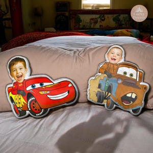 May include: Two custom-made car-shaped pillows featuring children's faces. One pillow is red, resembling Lightning McQueen, with the number 95. The other is a brown and blue tow truck, resembling Mater. Both pillows are on a bed.