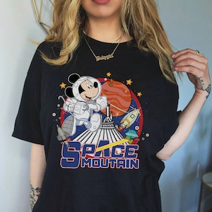 以下が含まれることがあります： スペースマウンテンの宇宙飛行士に扮したミッキーマウスのグラフィックがプリントされた黒いTシャツ。グラフィックには赤い惑星、星、ロケットが描かれている。グラフィックの下には「スペースマウンテン」の文字がプリントされている。