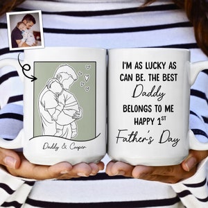 K&ouml;nnte beinhalten: Eine wei&szlig;e Keramiktasse mit einer schwarzen Linienzeichnung eines Vaters, der ein Kind h&auml;lt. Die Tasse tr&auml;gt den Text "Daddy & Cooper" und "I'm as lucky as can be. The best Daddy belongs to me Happy 1st Father's Day".
