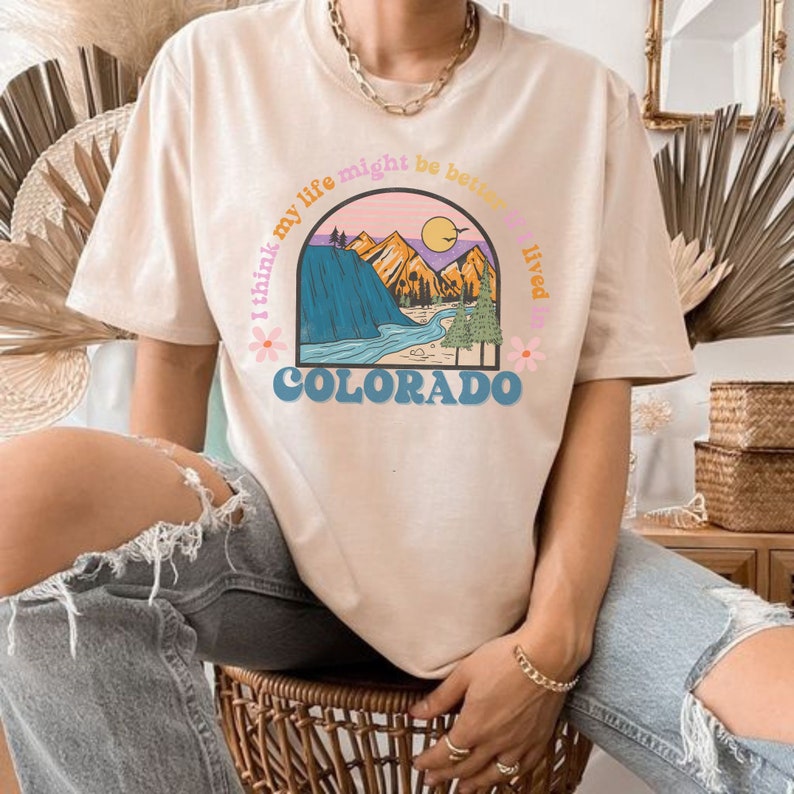 May include: A light beige t-shirt with a graphic print of a mountain range and a river. The text "I think my life might be better if lived in COLORADO" is printed around the graphic.