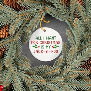 May include: White ceramic Christmas ornament with red and green holly and the text "All I want for Christmas is my Jack-a-poo" on a gray background.