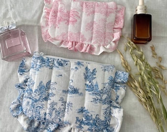 May include: Two toiletry bags with ruffled edges. One is pink with a tree design, the other is blue with a landscape scene. Both have a quilted texture and a zipper closure. The bags are on a white surface.