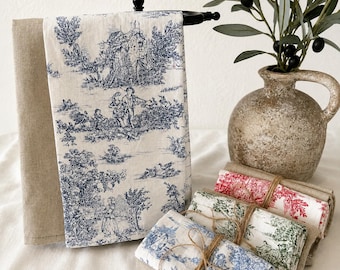 May include: A collection of linen kitchen towels with a blue and white toile pattern, displayed on a black towel rack. Additional folded towels in red, green, and blue toile patterns are bundled with twine. A beige vase holds an olive branch.