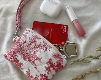 May include: A small, white and red toile print wristlet bag with a matching strap, a white AirPod case, a red debit card, a tube of lip gloss, and a key ring. The bag is open, revealing keys and a debit card.