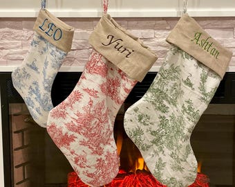 May include: Three Christmas stockings hang above a fireplace. Each stocking has a linen cuff with a name embroidered in a different color: blue, red, and green. The stockings are patterned with toile scenes in blue, red, and green.