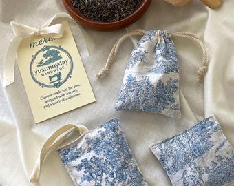 May include: Three small fabric pouches with a blue and white toile pattern filled with dried lavender. The pouches are on a white fabric background with a wooden bowl of lavender and a wooden scoop. A small card with the text "yusunnyday HANDMADE Custom-made just for you, wrapped with warmth and a touch of sunshine." is also on the background.
