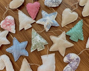 May include: A collection of handmade Christmas ornaments in various shapes, including stars, hearts, trees, candy canes, and stockings. The ornaments are crafted from fabric in a variety of patterns, such as gingham, stripes, and toile, and are strung together with twine.